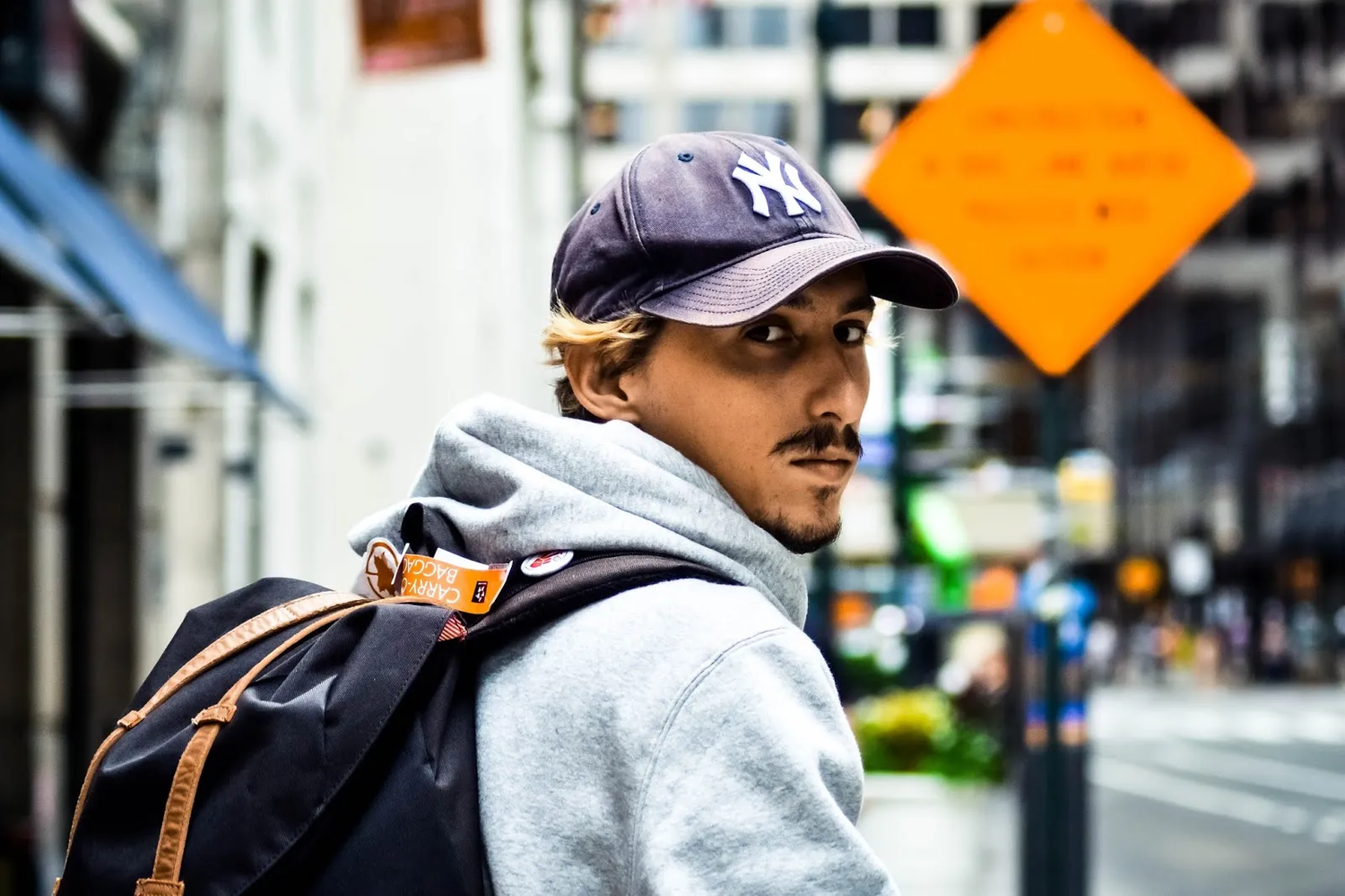 Portrait d’un jeune homme portant une casquette et un sweat à capuche, tourné vers l’objectif au milieu d’une rue animée.