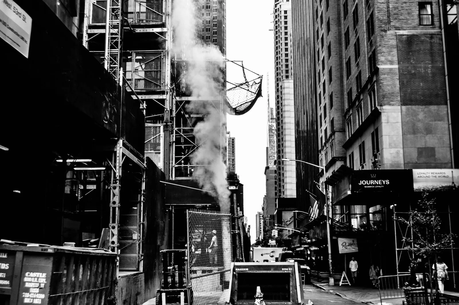 Scène urbaine en noir et blanc dans une rue étroite bordée de gratte-ciels, avec une colonne de vapeur s’échappant d’une bouche d’aération.