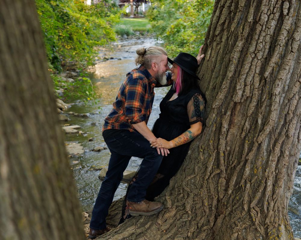 Couple in plaid and black clothing embrace by a large tree trunk near a creek in an outdoor nature setting.