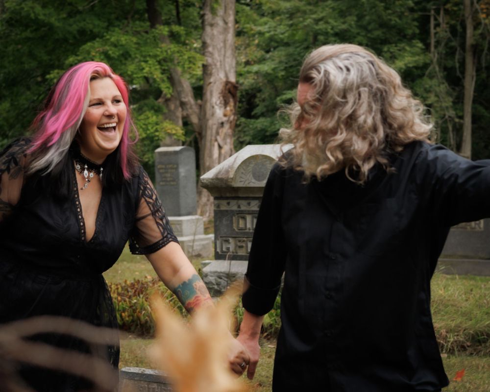 Two people in black outfits share a joyful moment together outdoors with visible laughter and pink hair details.