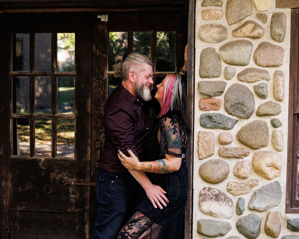 A couple embraces and laughs together against a rustic stone wall exterior next to a wooden door.