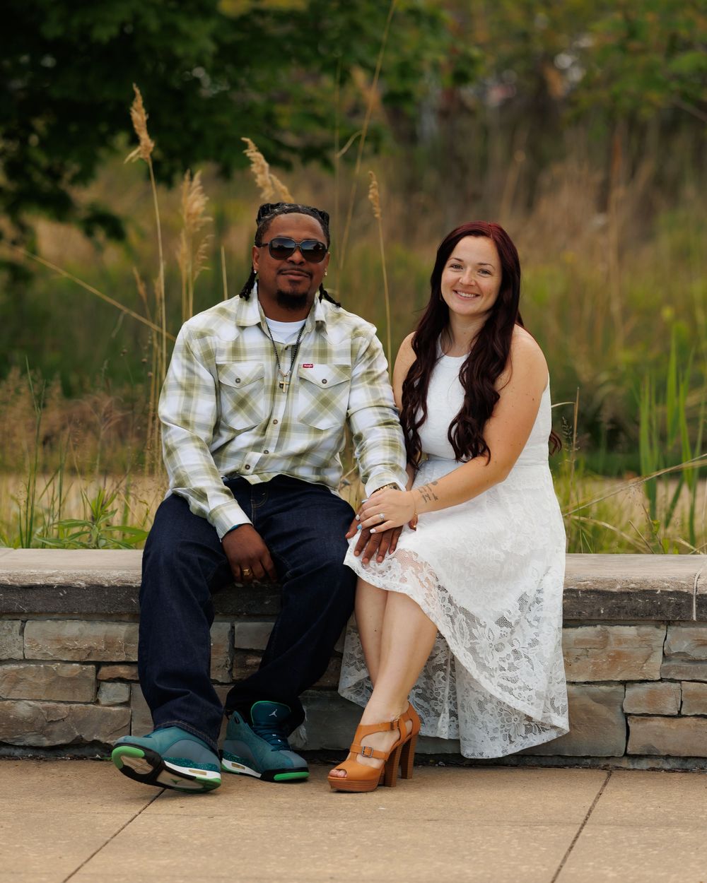 A couple sitting together on a stone wall outdoors, with tall grass and trees in the background.