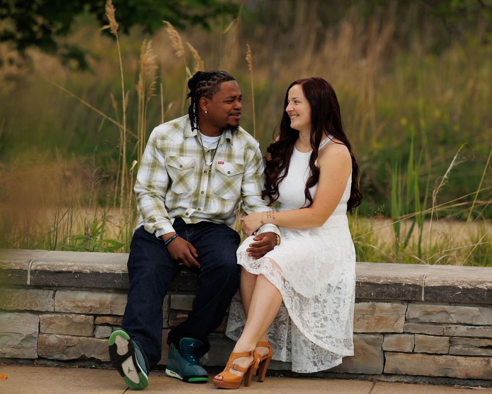 Two people share a tender moment while sitting on a stone wall surrounded by tall grass in a natural outdoor setting.