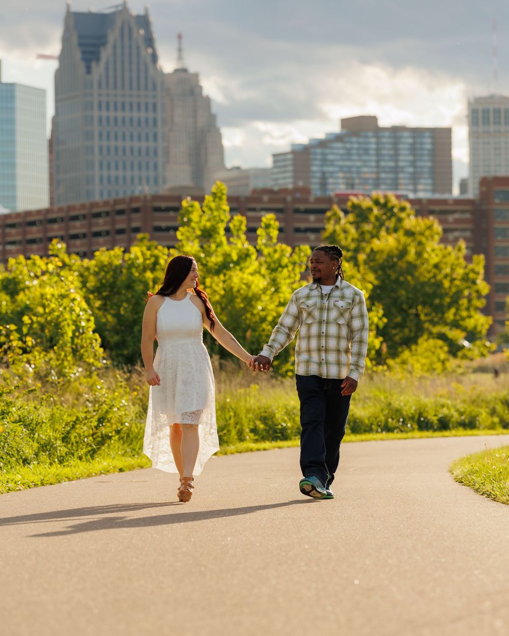 A couple walks together on a paved path with Detroit's downtown skyline and lush greenery in the background.