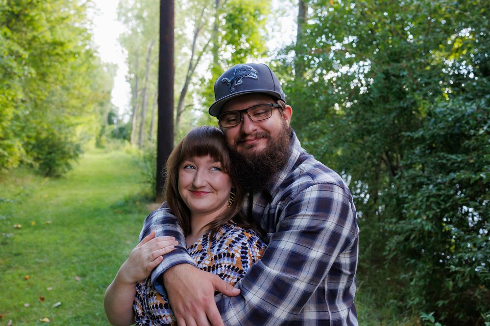 A couple in casual outdoor attire embraces while standing in a lush green park setting.