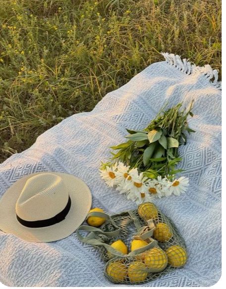 Picnic setup with white blanket, hat, flowers and lemons on grass.