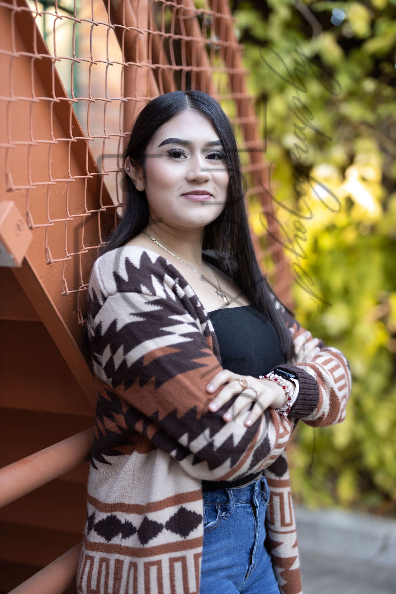 A person wearing a brown and beige patterned cardigan with geometric designs poses against an orange wall with foliage in background.