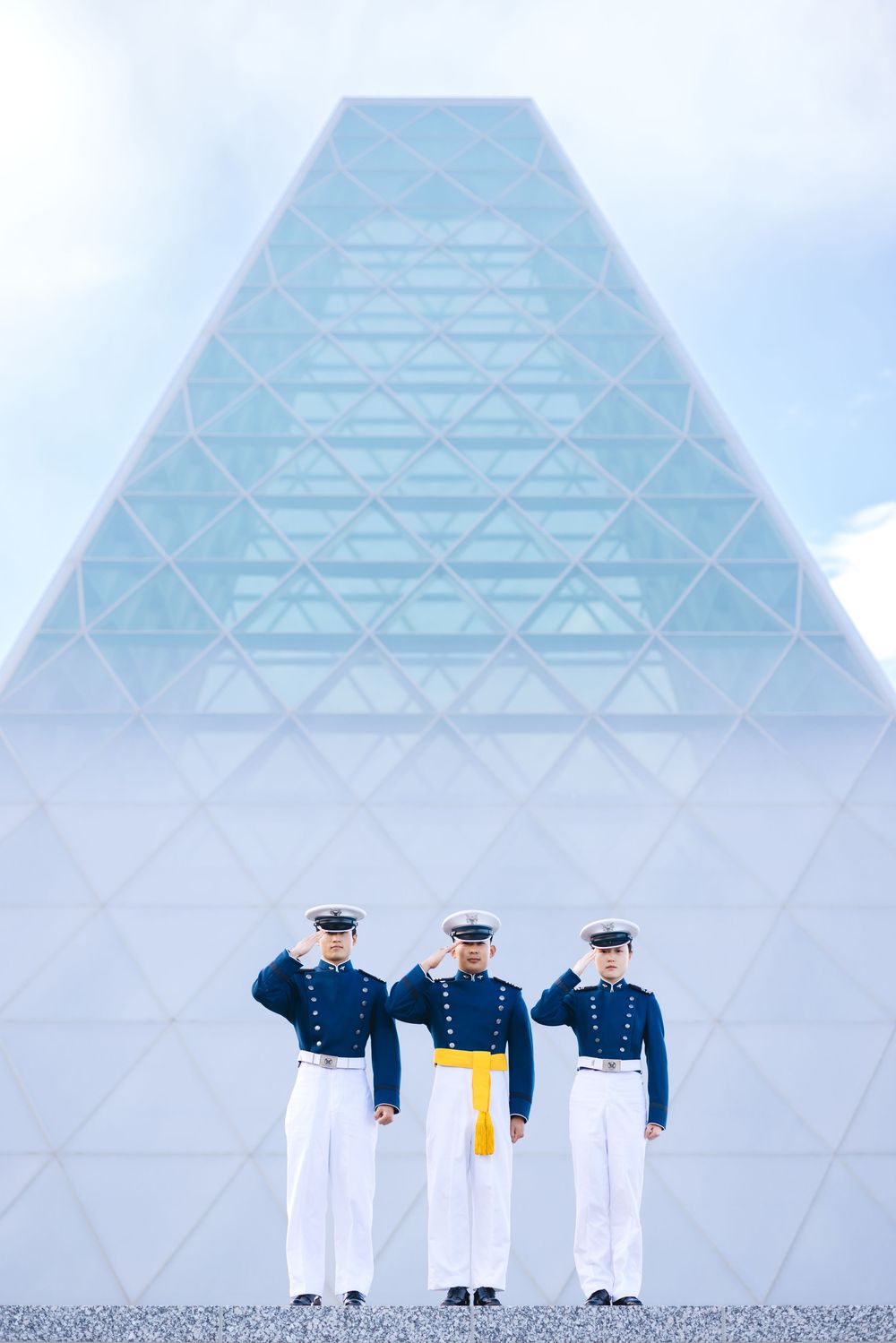 Three military cadets in dress uniforms saluting before a modern glass pyramid building under cloudy skies.