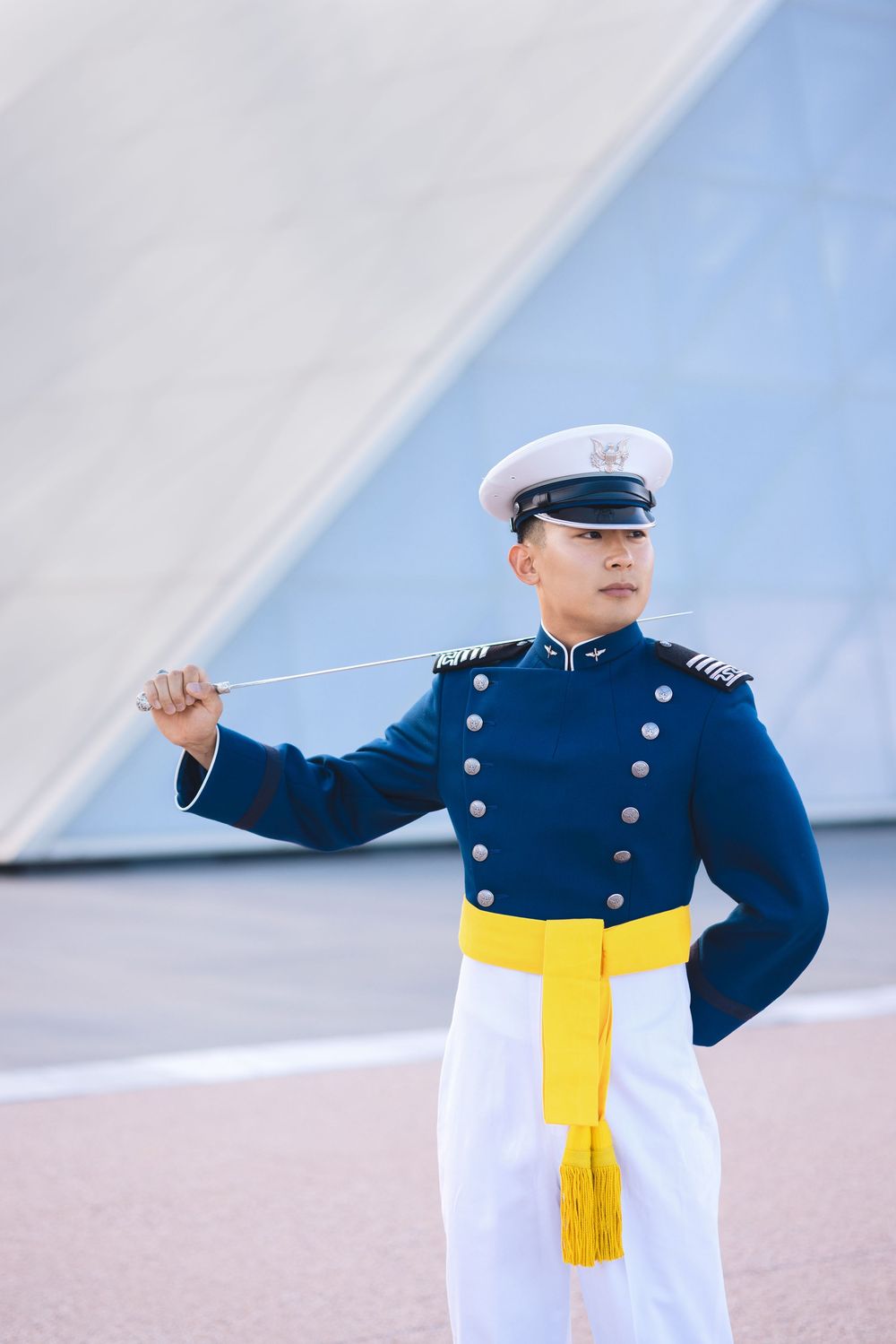 Military officer in blue dress uniform with gold sash and white cap holds a sword outdoors.