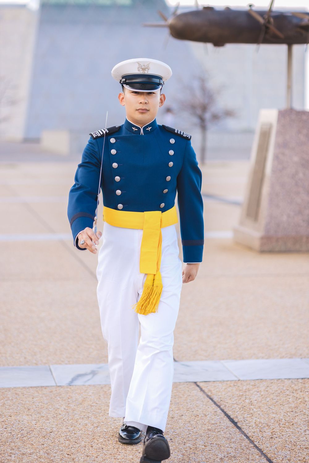 Military cadet in blue dress uniform with gold sash and white cap marching outdoors.