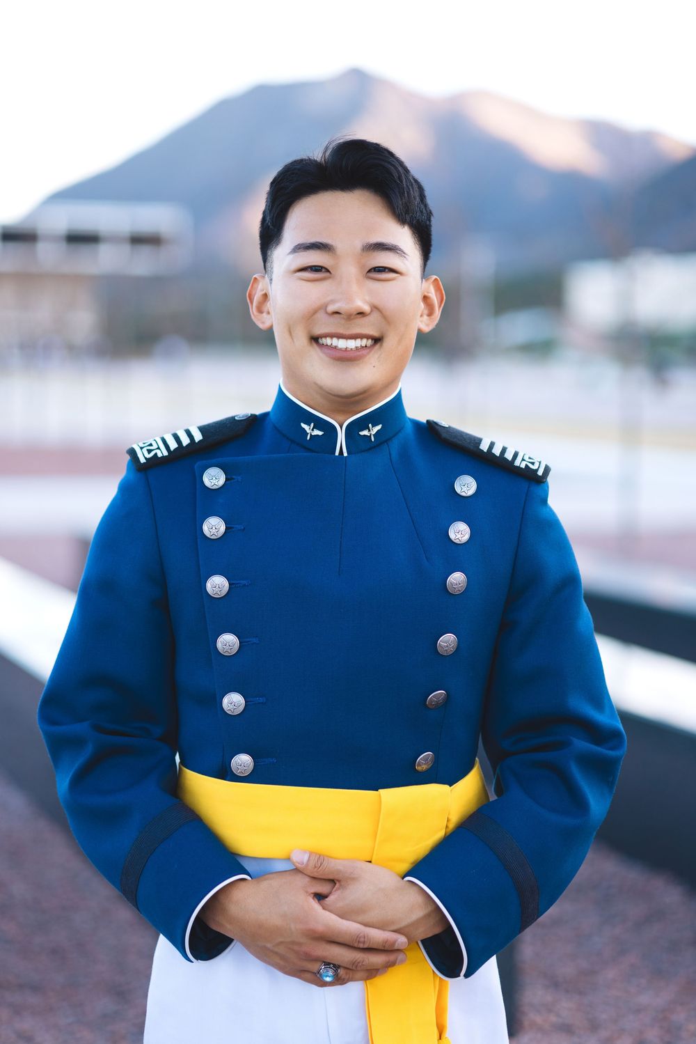Smiling young man in blue military dress uniform with yellow sash and silver buttons outdoors.