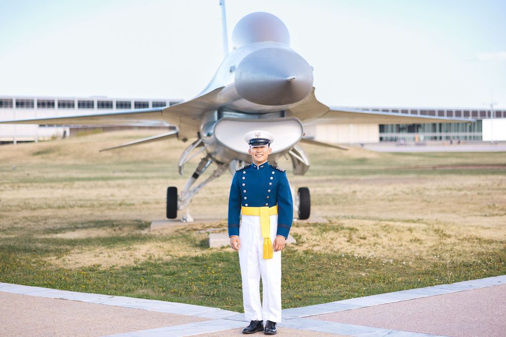 Military cadet in dress uniform stands before an F-16 fighter jet at the US Air Force Academy.