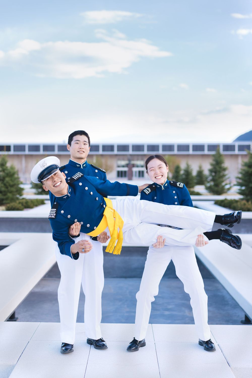 Three military cadets in blue dress uniforms playfully lifting a fellow cadet outdoors.