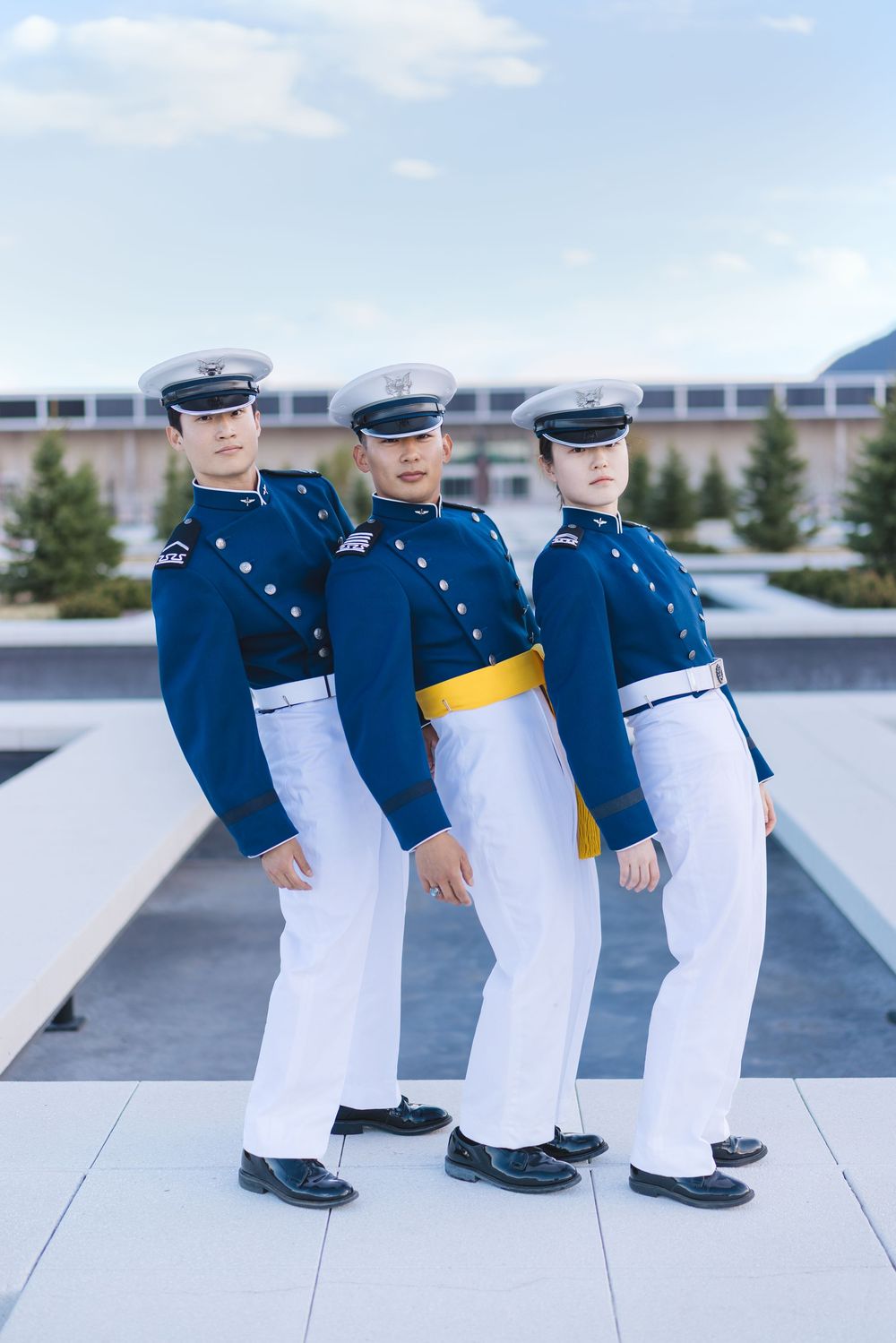 Three military cadets in blue dress uniforms with white pants and caps standing at attention outdoors.