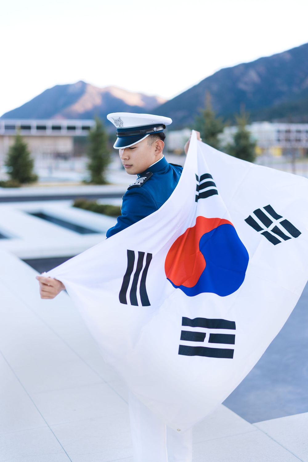 South Korean military officer in uniform holding a large Taegukgi flag outdoors with mountains in the background.