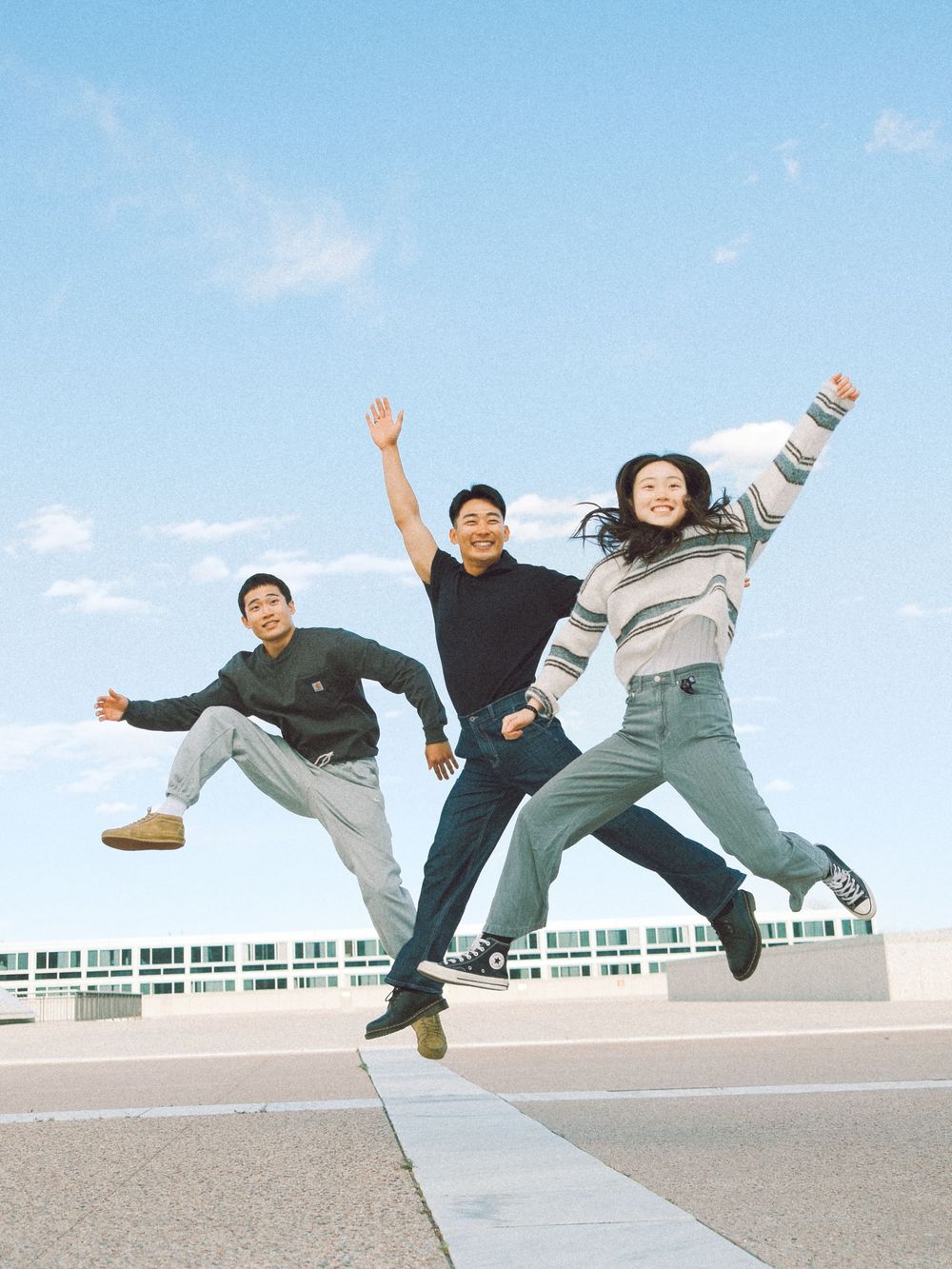 Three young Asian people joyfully jumping in the air outdoors against a blue sky.