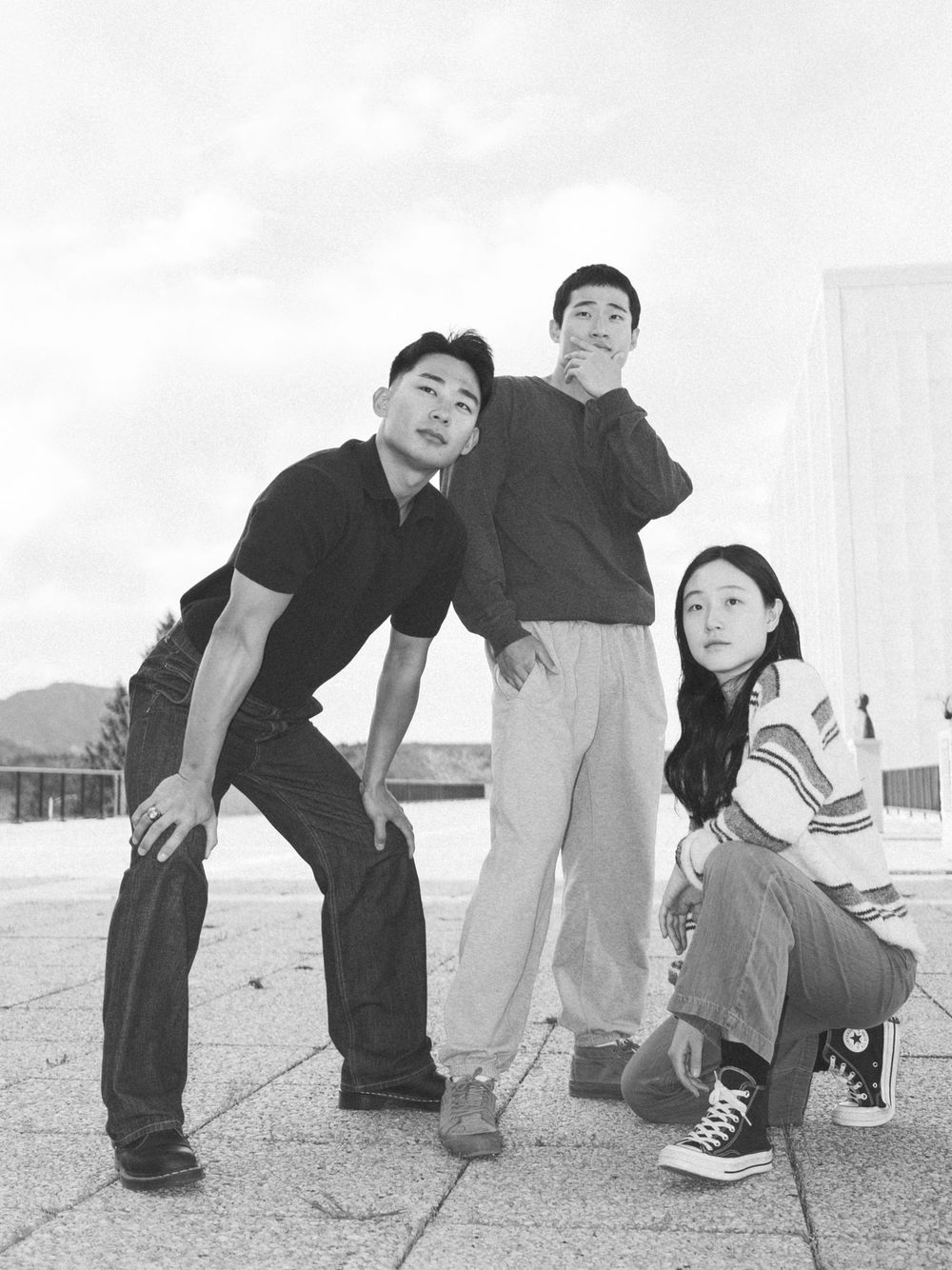 Three young people pose outdoors in a black and white photo, two standing and one crouching, wearing casual streetwear.