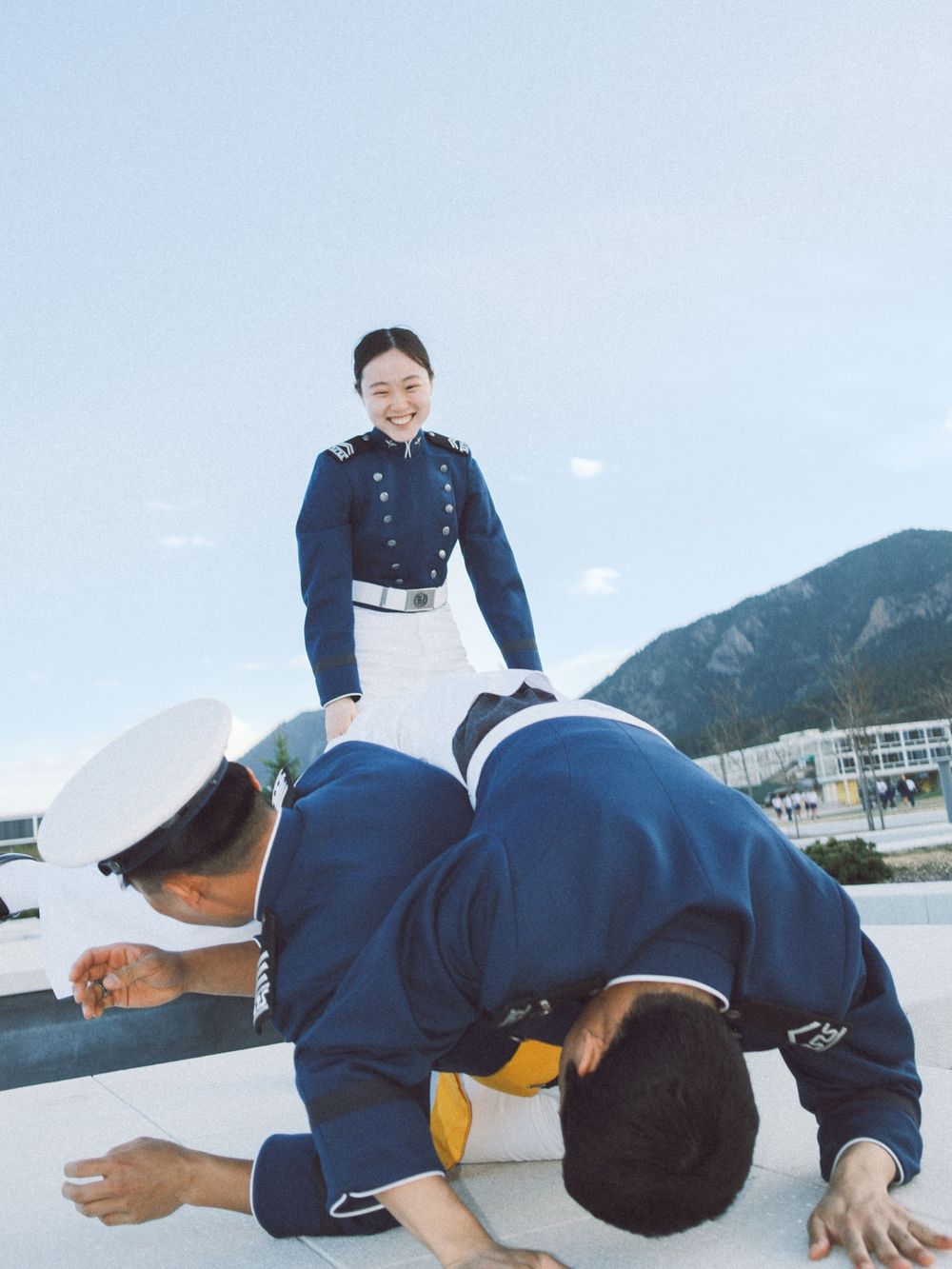 Smiling young woman in military uniform standing while two cadets bow before her outdoors.