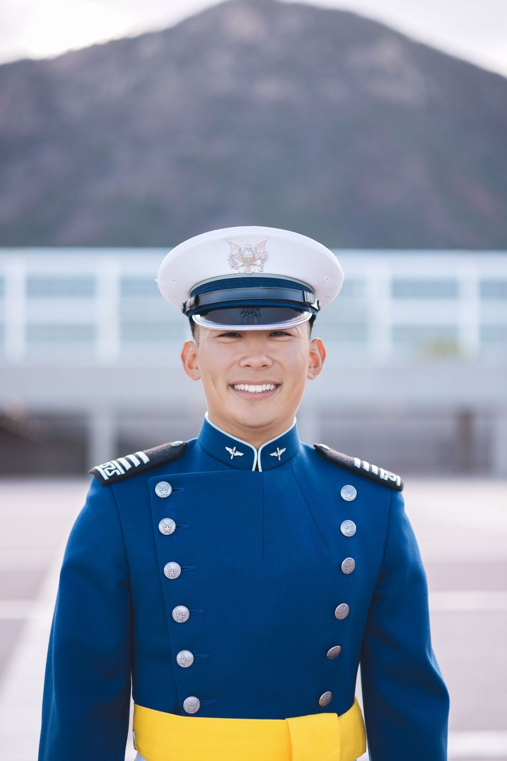 Smiling cadet in blue military dress uniform with white cap and gold trim stands outdoors near mountain backdrop.