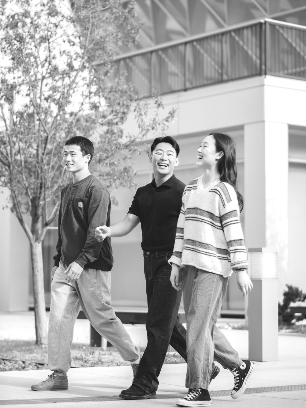 Three young Asian people laughing and walking together outdoors near a modern building in black and white photo.