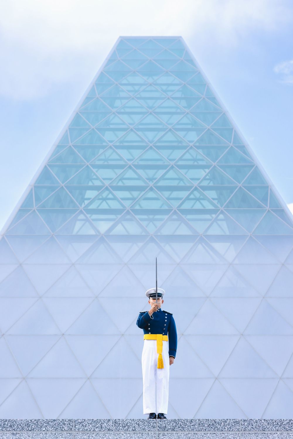 Military guard in dress uniform stands at attention before a large glass pyramid building under a cloudy sky.