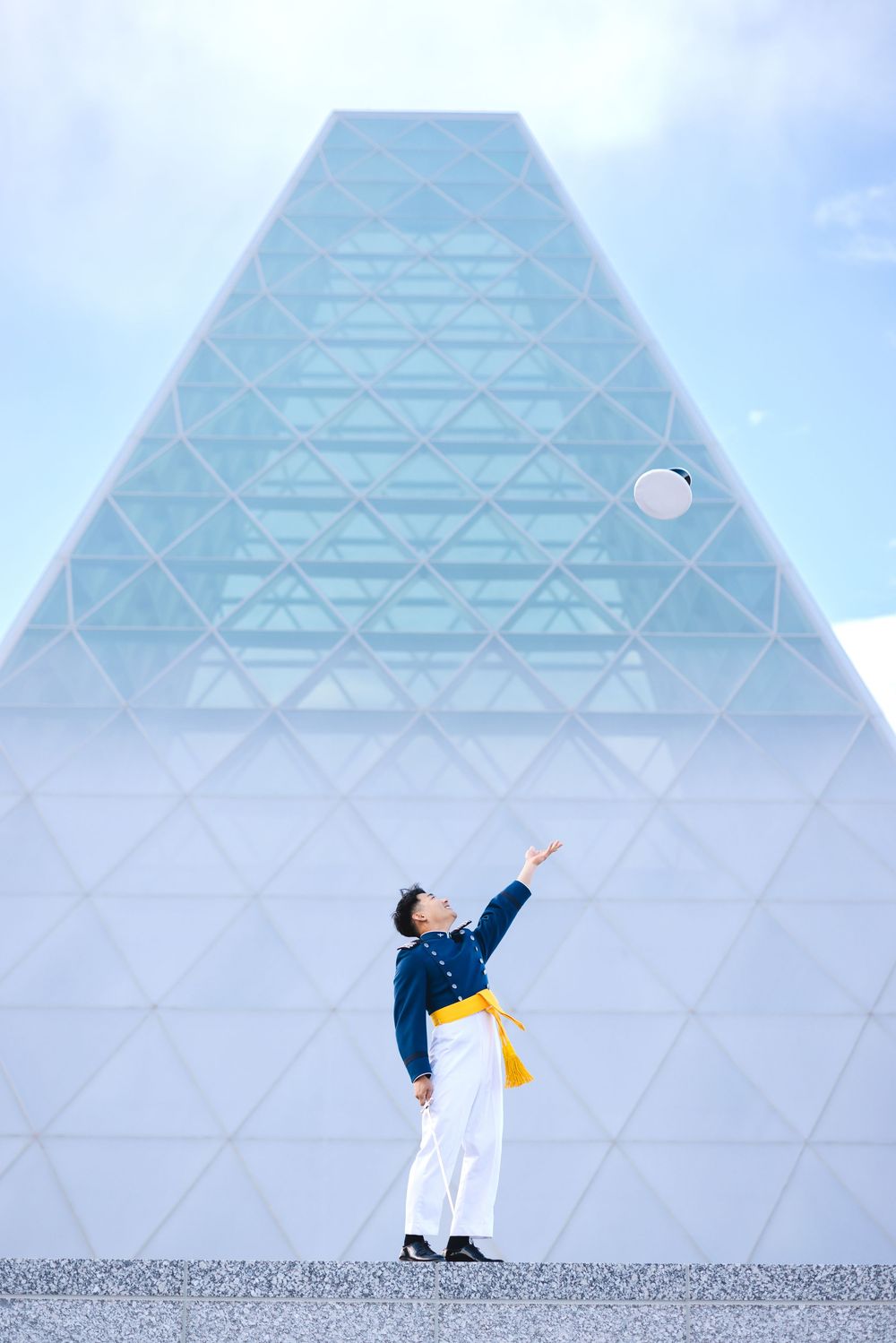 Man in blue military uniform tossing white hat in front of modern glass pyramid building.