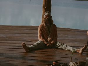 Person in brown clothing sits cross-legged on wooden deck in meditative pose.