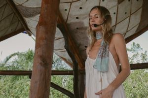Person in flowy white garment poses under wooden gazebo structure with greenery in background.