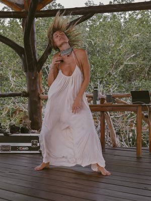 Figure in flowing white dress dances freely on wooden deck surrounded by trees and nature.