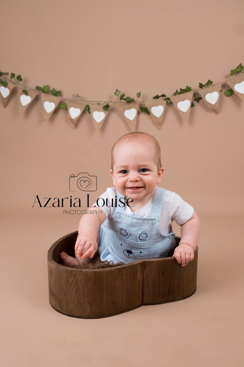Baby wearing light denim overalls sits in wooden heart-shaped bowl under heart banner garland on beige backdrop.