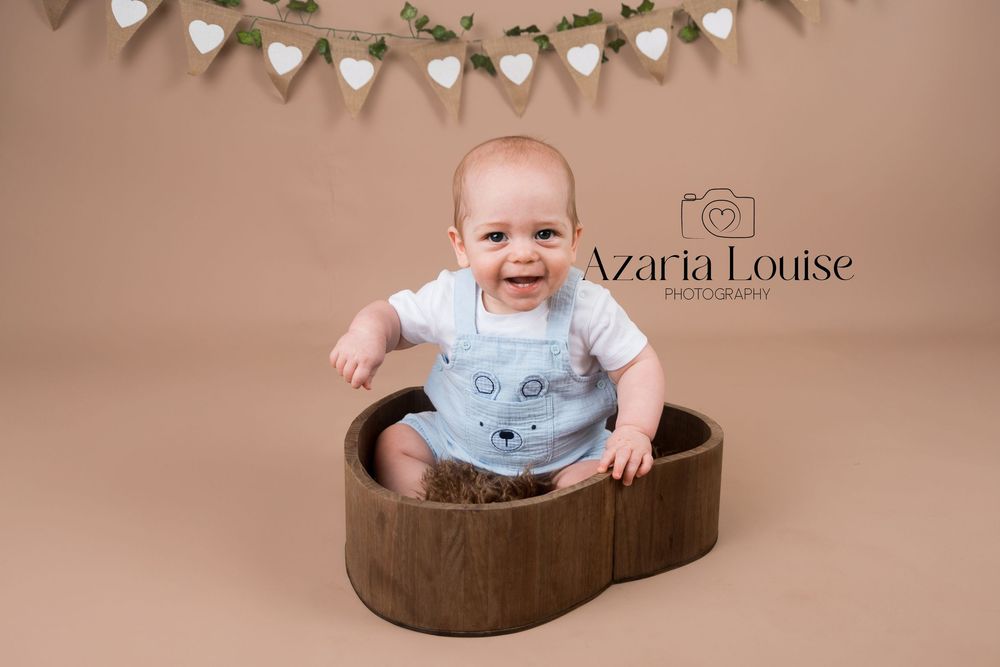 Smiling baby in light blue outfit sits in heart-shaped wooden prop with bunting banner hanging above on beige backdrop.