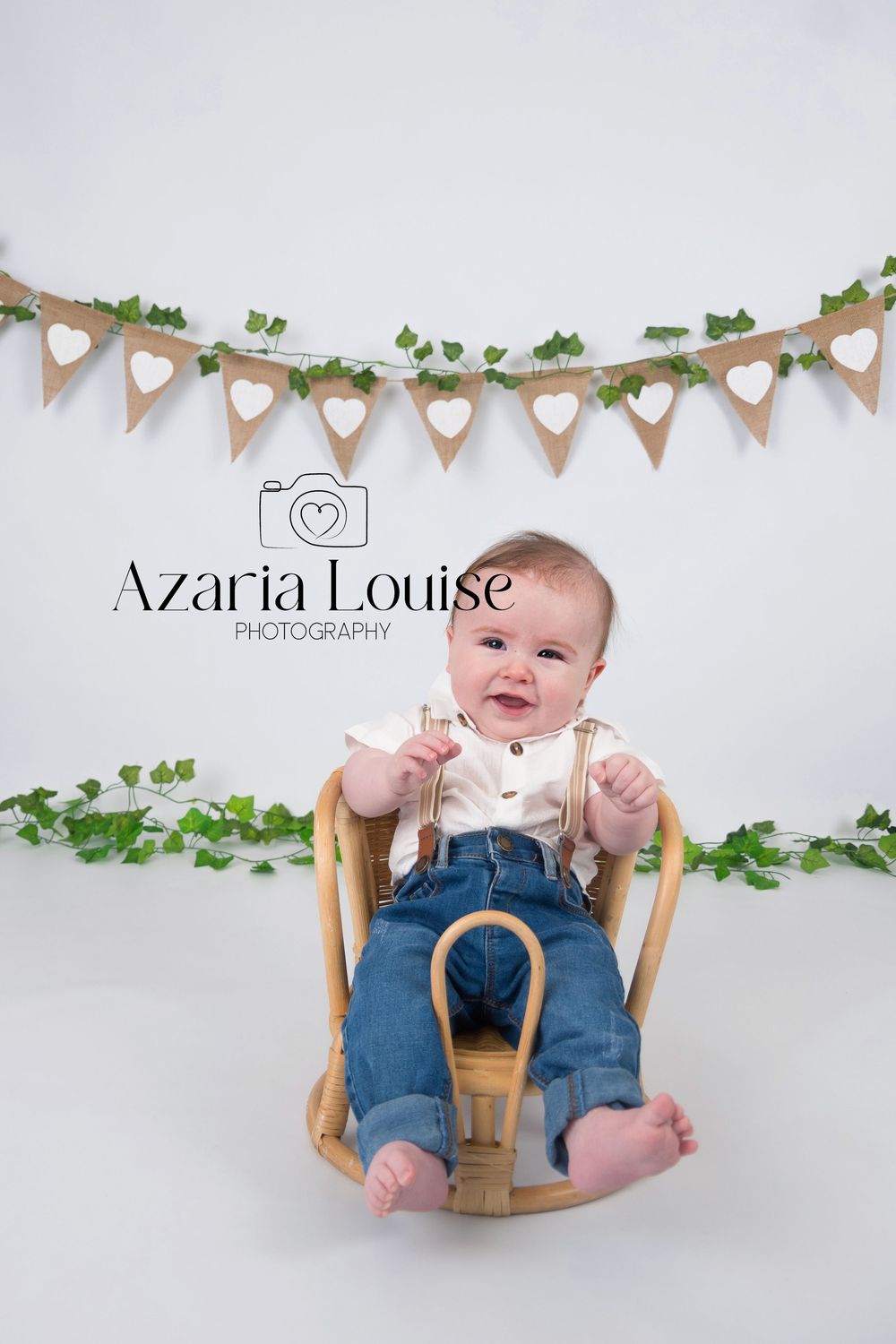 Baby sitting in wooden rocking chair with heart banner garland and greenery decoration on white backdrop.