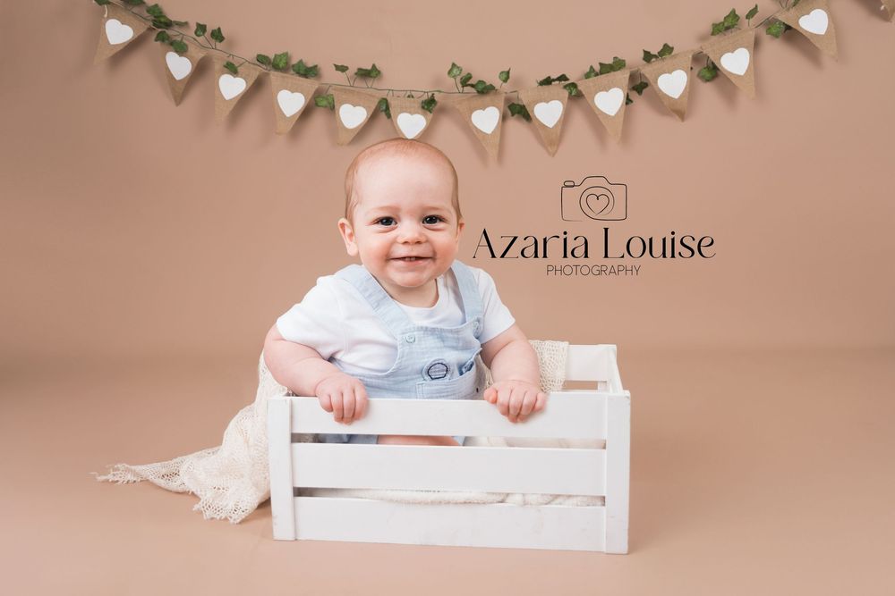 Baby sits in white wooden crate with heart banner decoration against beige backdrop in professional studio photo.
