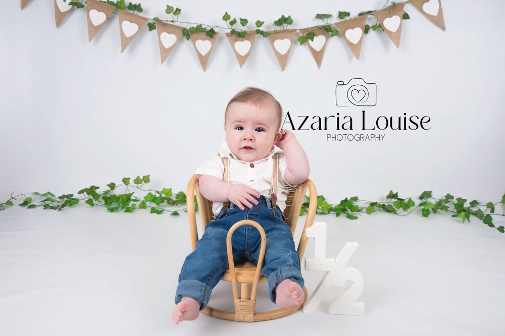 Baby sits in wooden rocking chair under bunting banner with vine decorations against white backdrop.