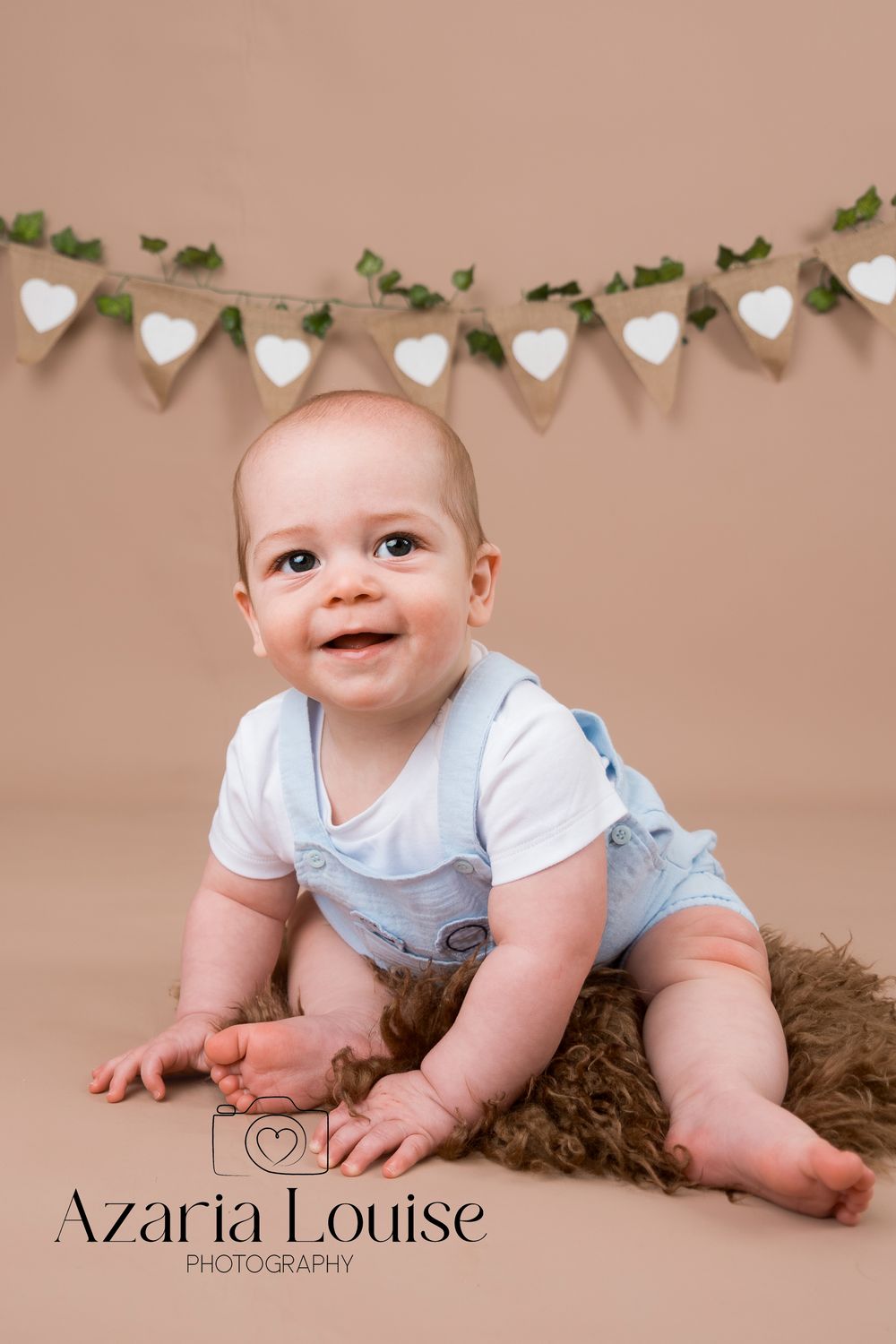 Baby wearing blue overalls crawls on brown blanket under heart garland decoration against beige backdrop.