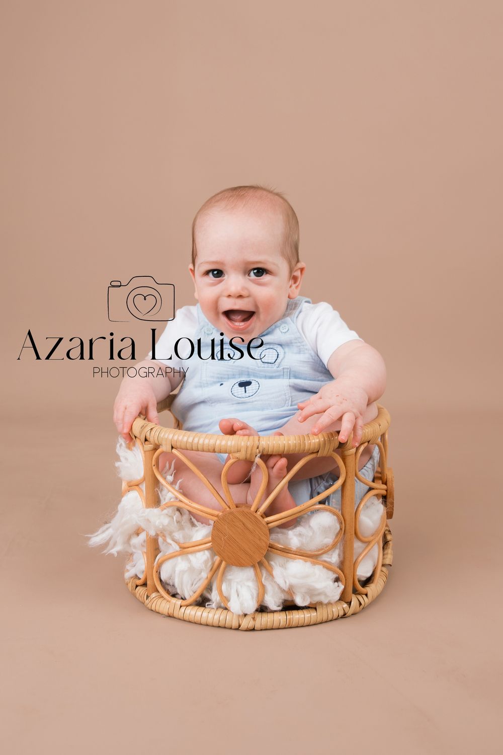 Smiling baby wearing a white shirt sits in a decorative woven basket with cotton accents against a neutral background.