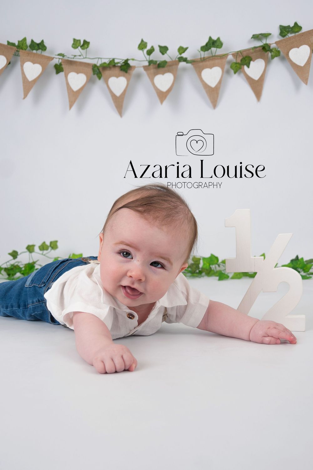 A white bunting banner decorated with hearts hangs above a baby lying on a white backdrop with greenery accents.
