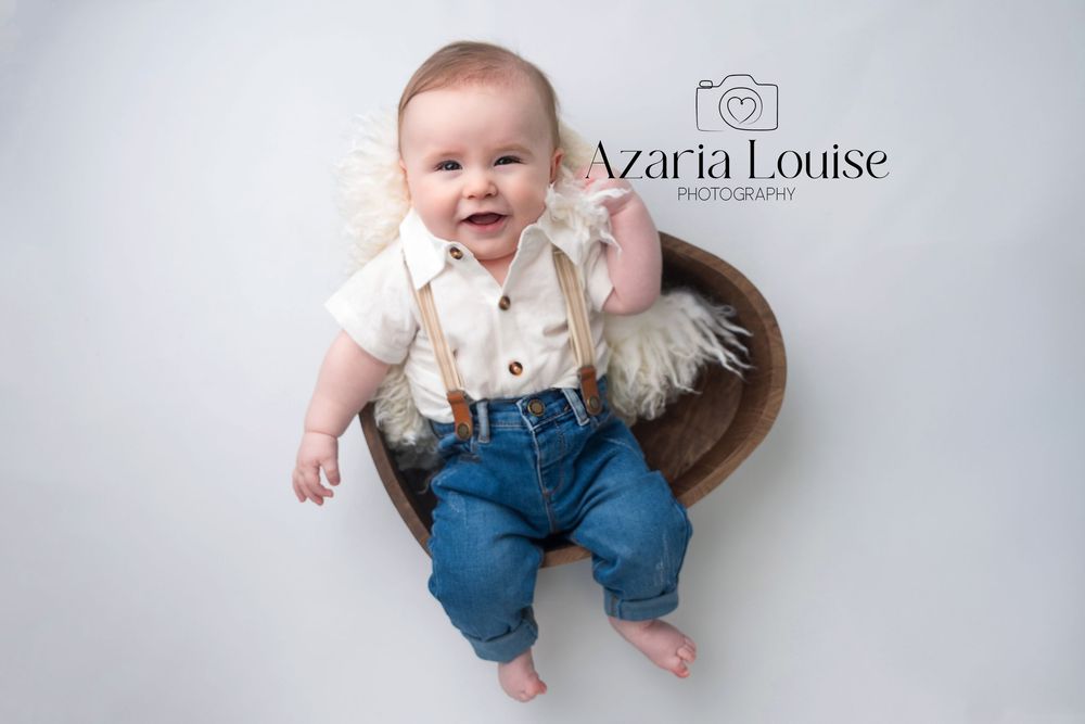 A joyful baby wearing a white shirt with suspenders and blue pants sits in a wooden bowl on a light gray backdrop.