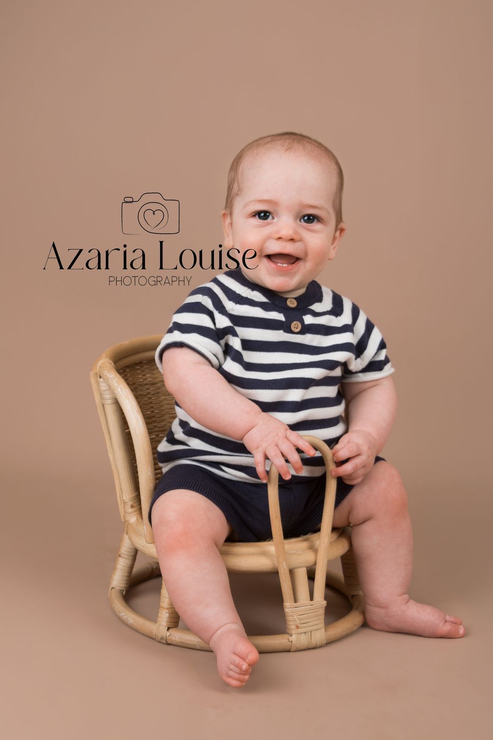 A baby wearing a striped polo shirt sits smiling in a small wicker chair against a brown backdrop.