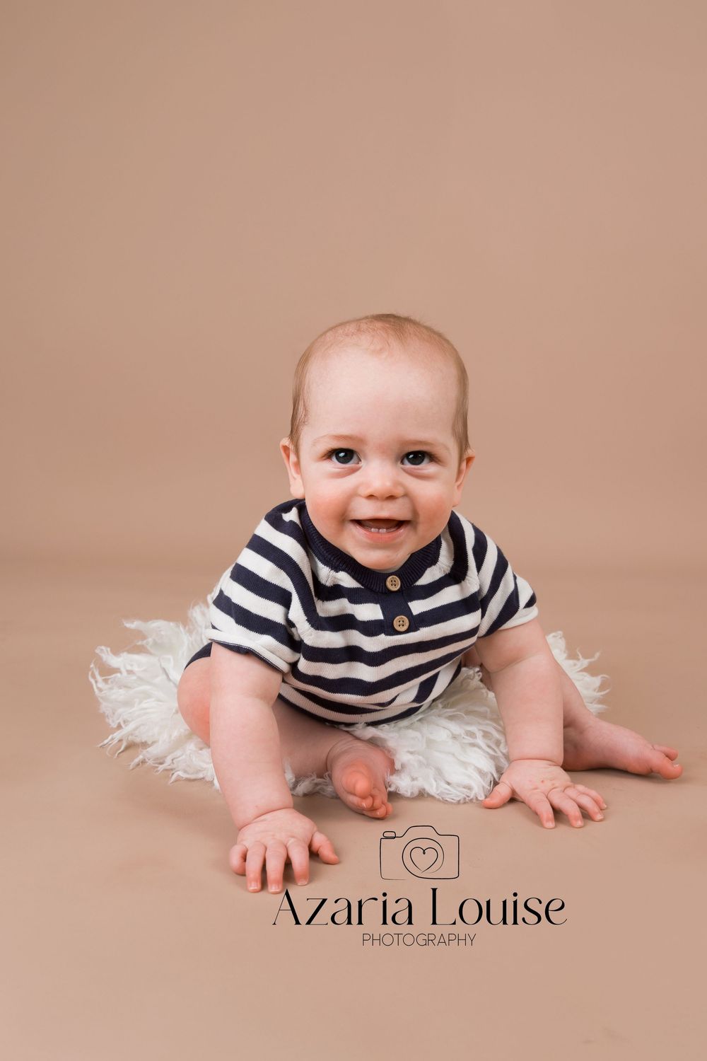 Baby wearing black and white striped shirt sits on white fur rug during professional photo session.