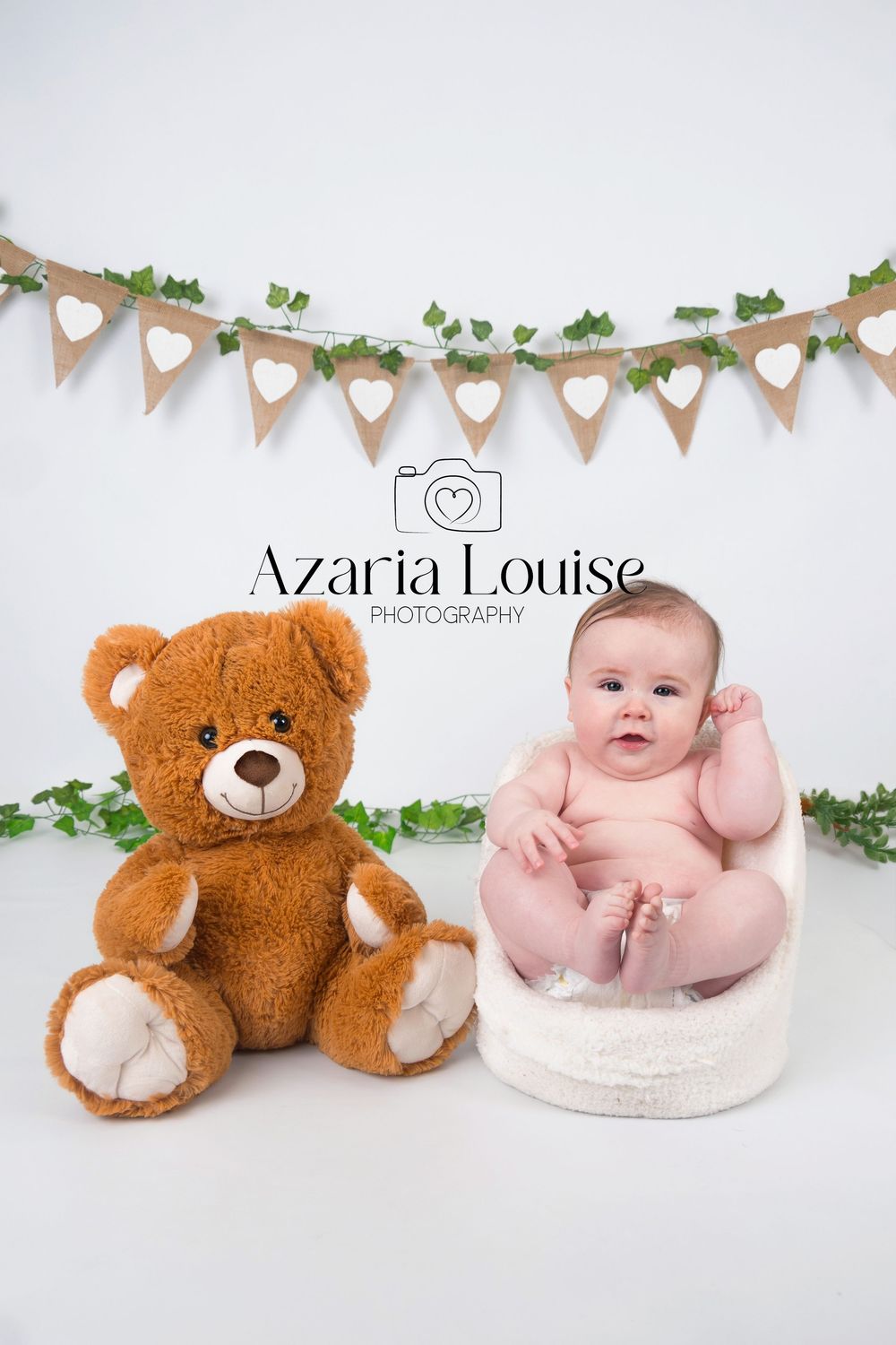 Baby sitting on white cushion next to brown teddy bear with heart banner garland decoration above.