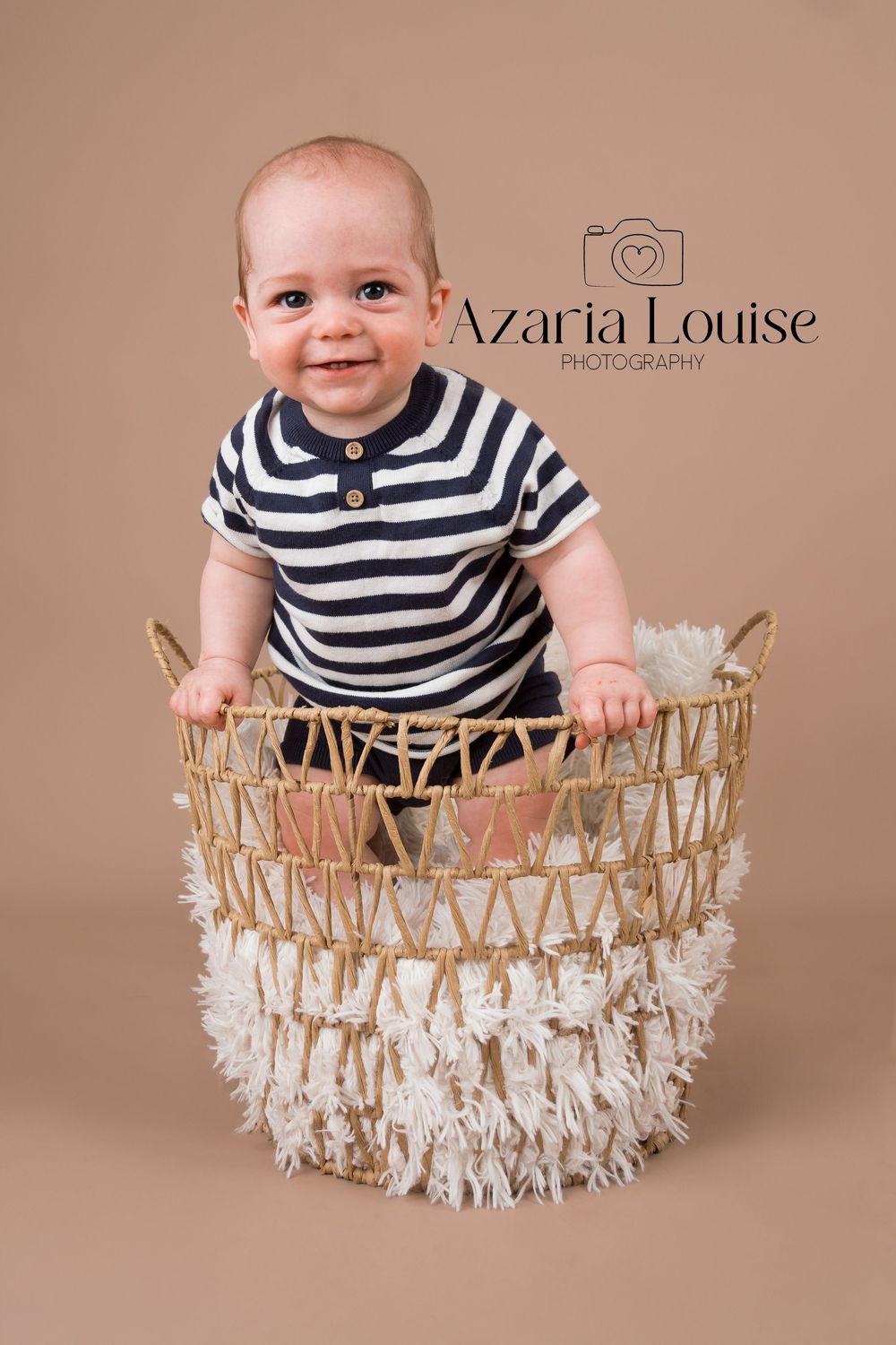 Baby in striped navy shirt sits in decorative woven basket with macrame fringe against brown backdrop.