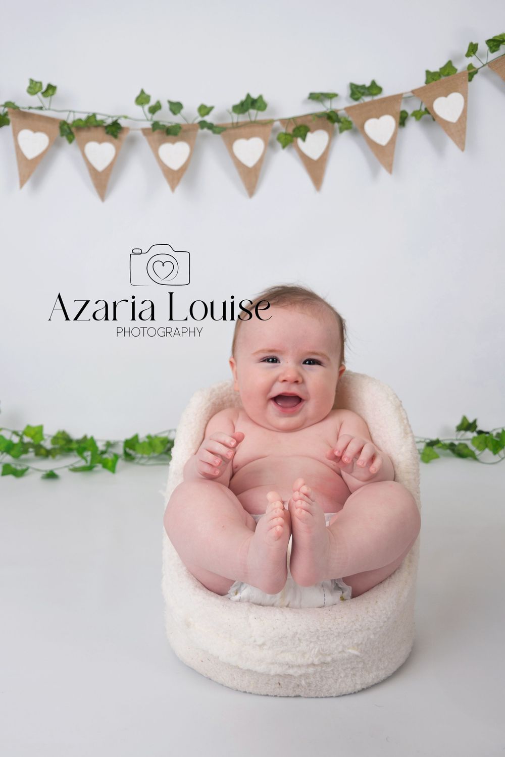 Professional baby photography with rustic burlap heart banner and ivy garland decorations against white backdrop.
