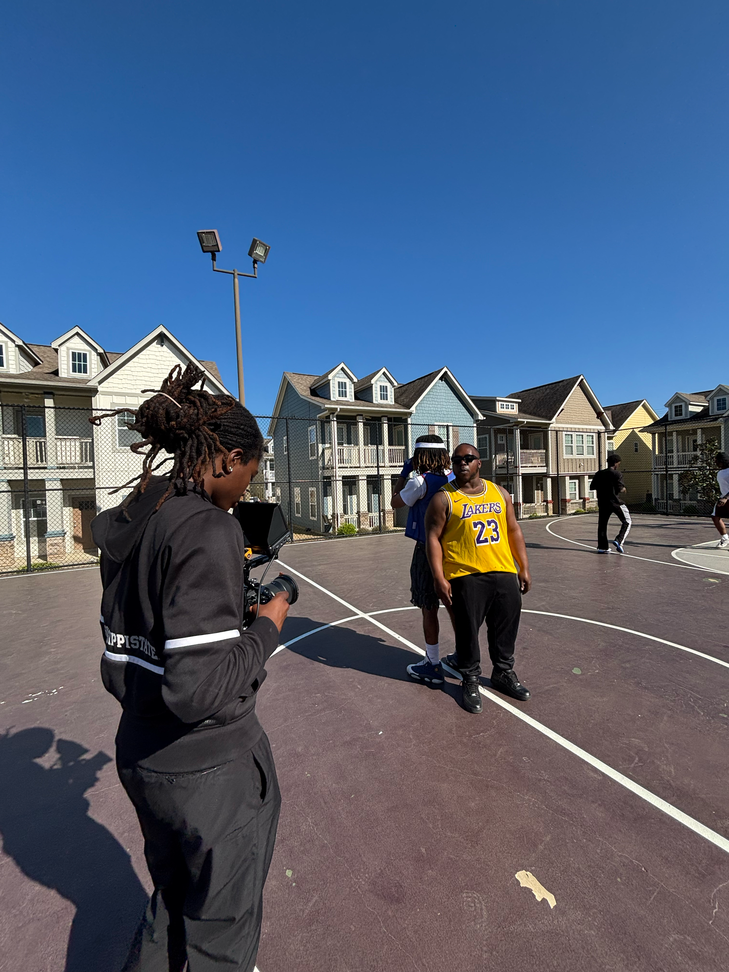 Mills filming two individuals in Lakers jersey #23 on outdoor basketball court with residential townhouses in background.