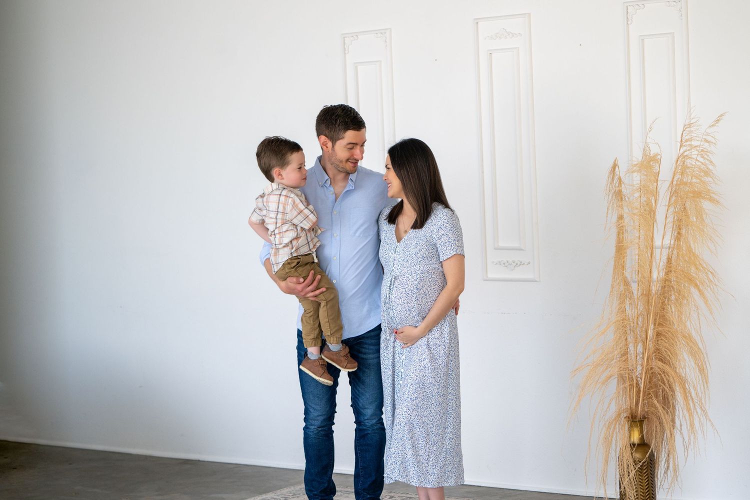 A family stands together in a minimalist white room decorated with pampas grass in the corner.