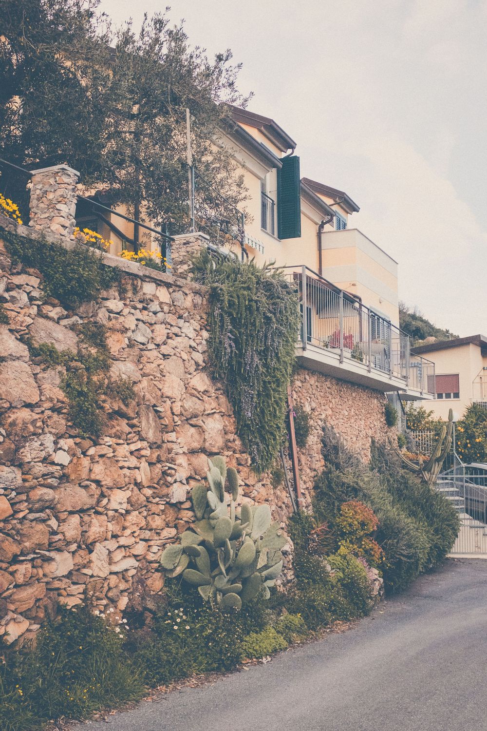 A rustic stone wall lines a narrow street with Mediterranean-style houses and cascading plants in a quaint village setting.