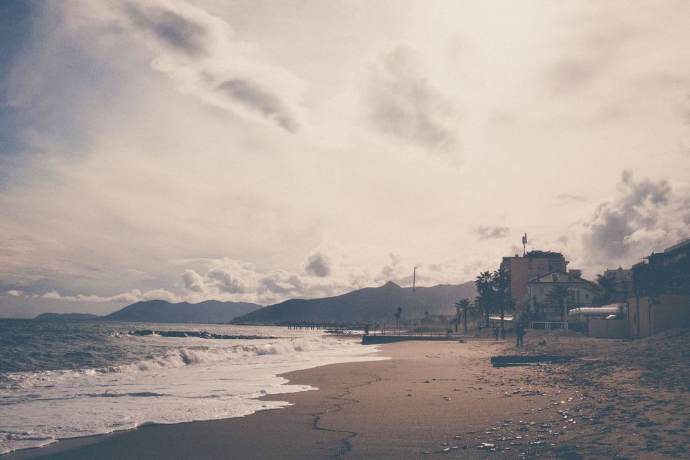 A moody coastal beach scene at sunset with wind turbines and buildings silhouetted against cloudy skies.