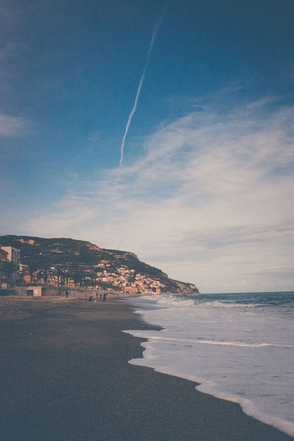 A scenic Mediterranean coastline with cliffs and buildings nestled on hillside under blue sky with white contrail.