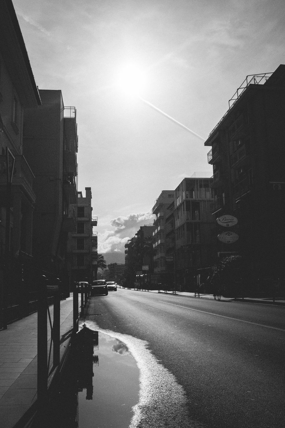 A black and white urban street scene with tall buildings and dramatic sunlight reflecting off a wet road surface.