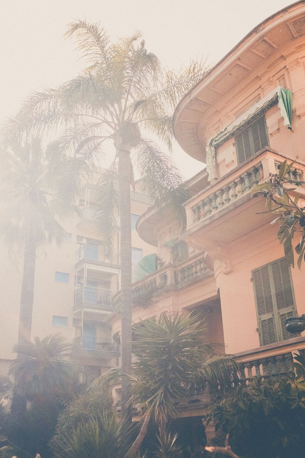 A pink Mediterranean-style building with palm trees and tropical plants against a misty or hazy sky.